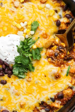 A cast iron skillet filled with a quinoa enchilada bake with sour cream and cilantro on top with two spoons sticking out.