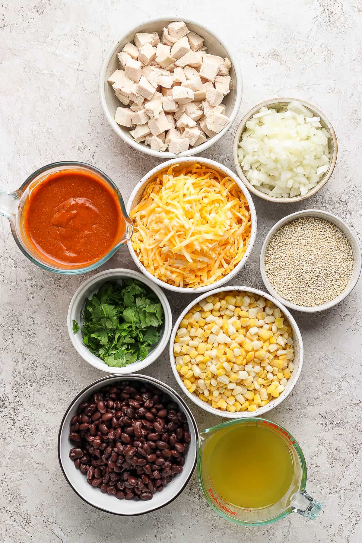 Individual bowls of chicken, diced onion, shredded cheese, enchilada sauce, quinoa, cilantro, corn, balck beans, chicken stock.