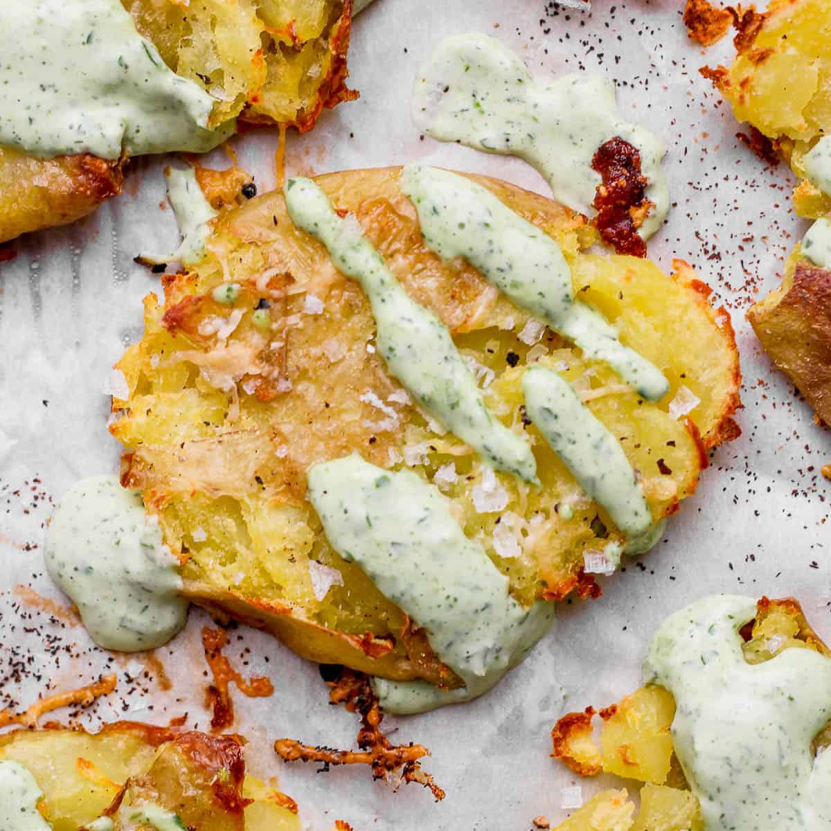 A smashed potato on piece of parchment with herby green sauce drizzled over top.