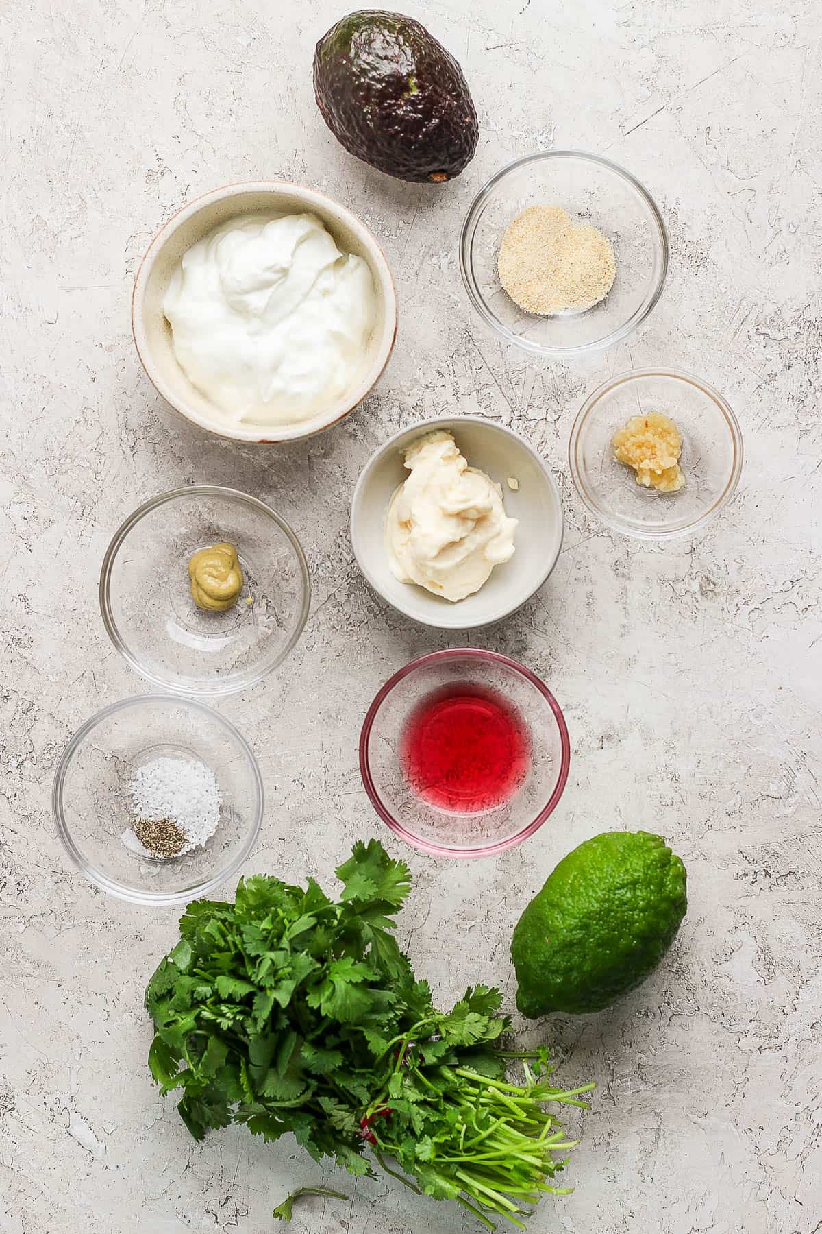 Individual bowls of mayo, minced garlic, greek yogurt, dijon mustard, salt, a whole avocado, a bunch of cilantro, a full lime, ground pepper, red wine vinegar, lime juice, and garlic powder.