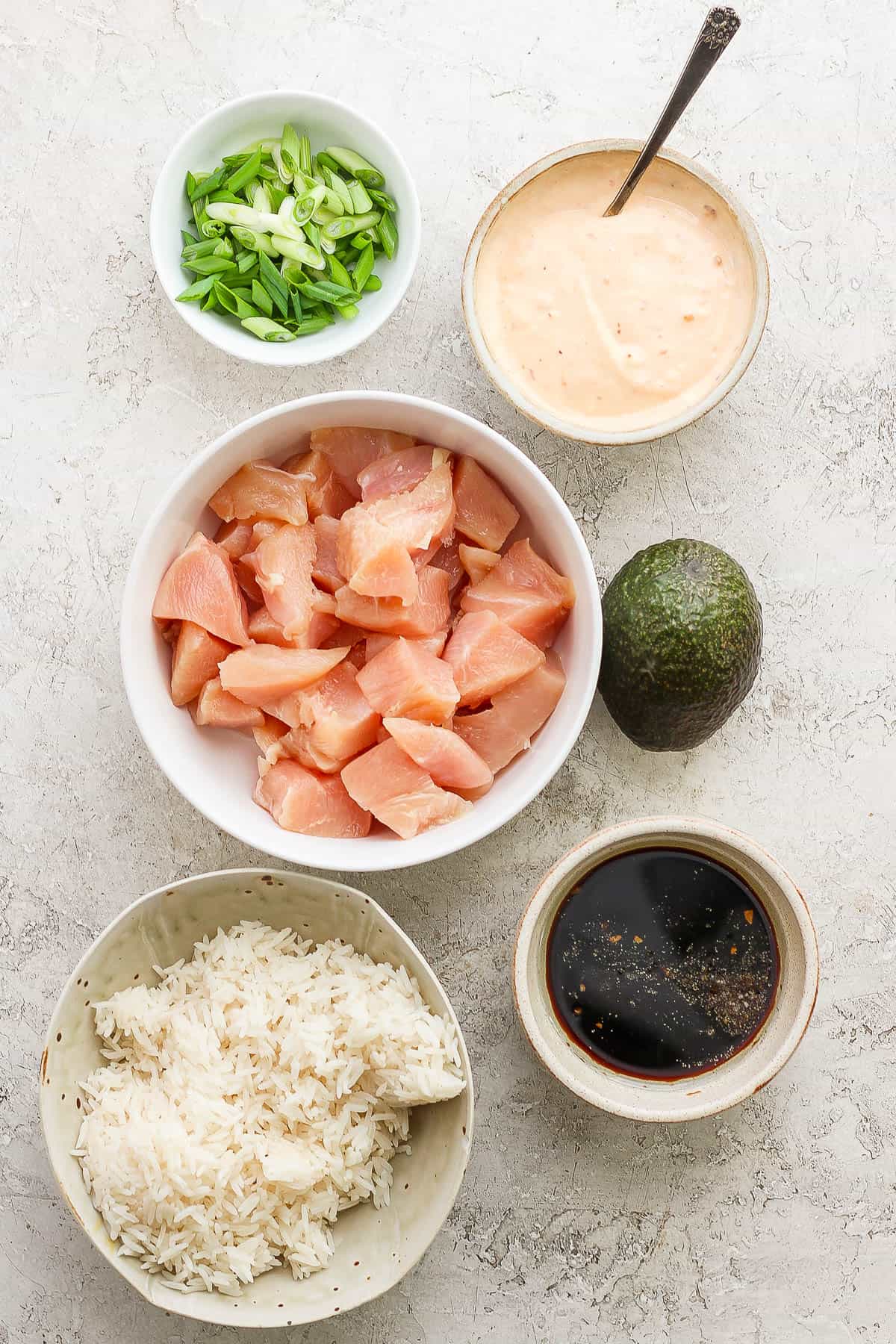 Individual bowls of cubed chicken, sliced green onions, cooked white rice, bang bang sauce, chicken marinade, and a whole avocado.