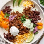 A beef taco rice bowl with ground taco meat, tomatoes, onion, shredded cheese, sour cream and fresh lime wedges over rice.