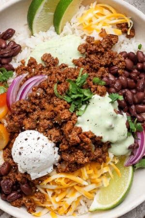 A beef taco rice bowl with ground taco meat, tomatoes, onion, shredded cheese, sour cream and fresh lime wedges over rice.