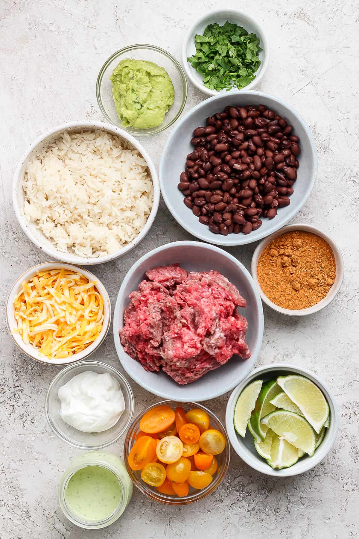 Individual bowls of chopped cilantro, guacamole, black beans, white rice, seasonings, shredded colby jack cheese, ground beef, sour cream, lime wedges, halved cherry tomatoes, and avocado lime ranch dressing.