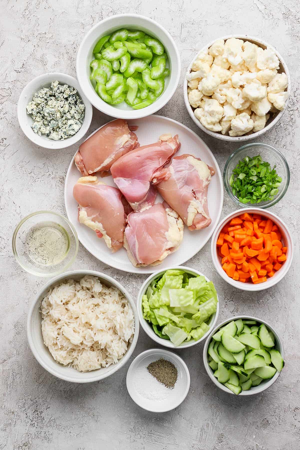 Individual bowls of sliced celery, blue cheese, cauliflower, chicken thighs, green onions, carrots, oil, romaine lettuce, cooked rice, salt & pepper, sliced and cut cucumbers.