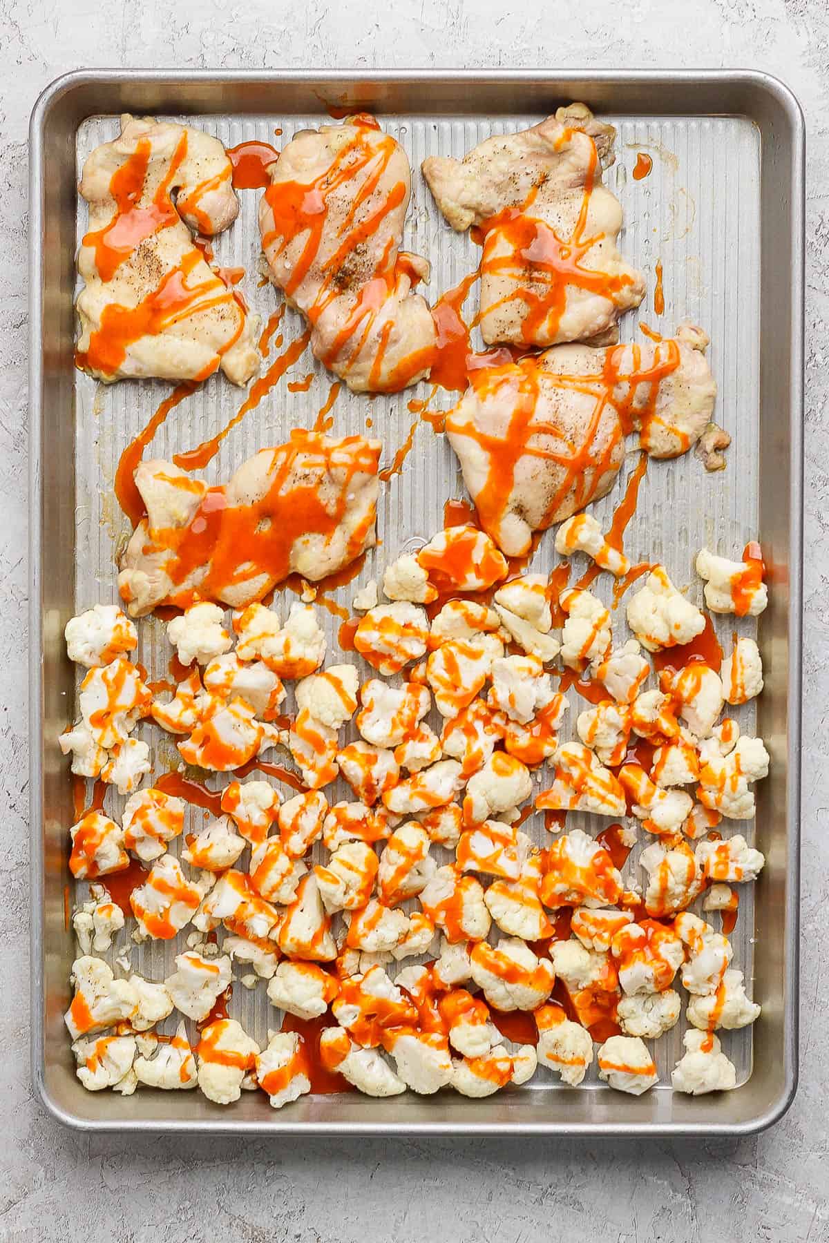 Baked chicken thighs and roasted cauliflower on a baking sheet drizzled with buffalo sauce to make a buffalo chicken rice bowl.