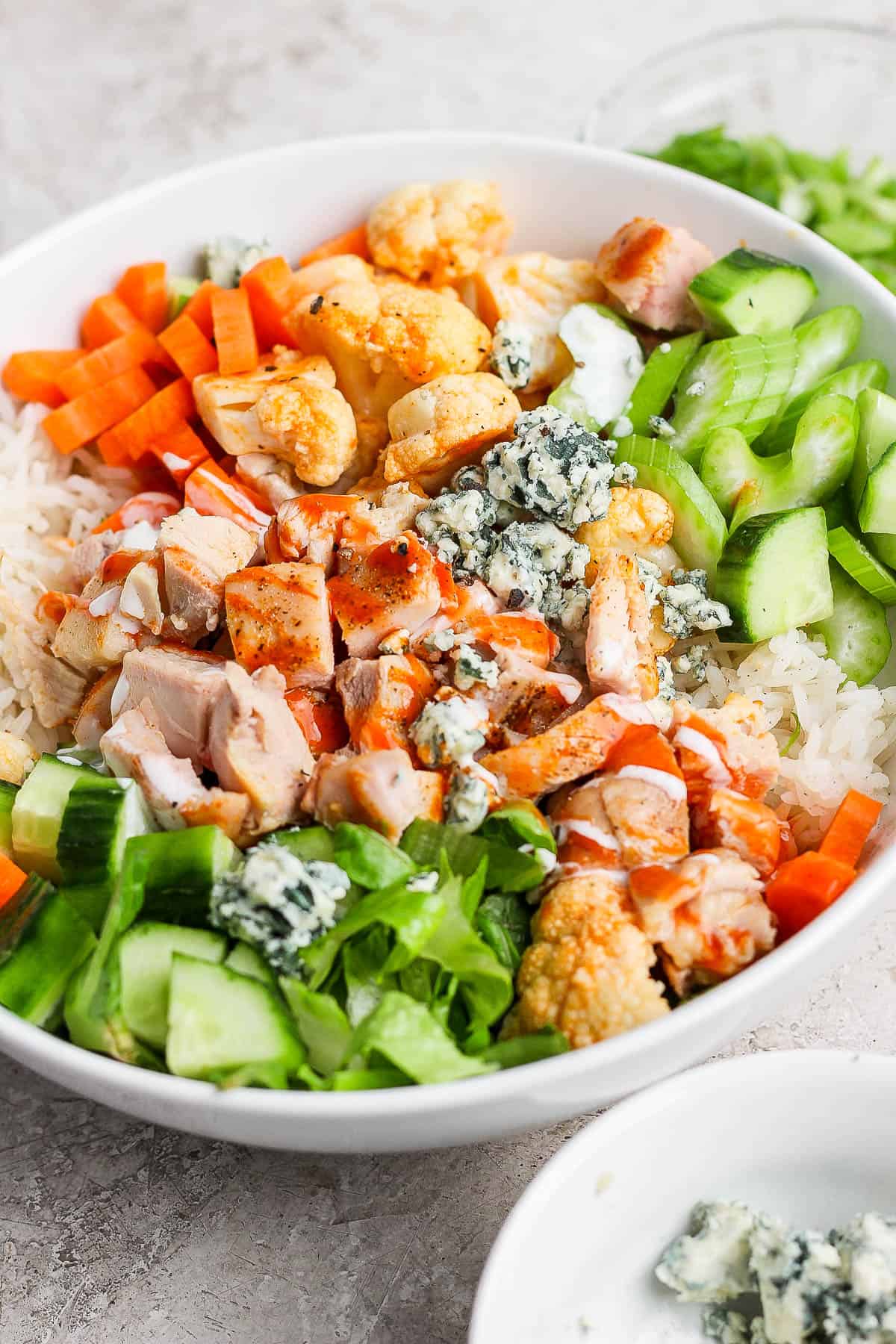 A bowl with rice, carrots, cucumber, celery, blue cheese, cubed chicken and buffalo sauce and ranch.