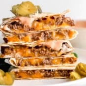 A stack of cheeseburger quesadillas with pickles and burger sauce on top and next to it.