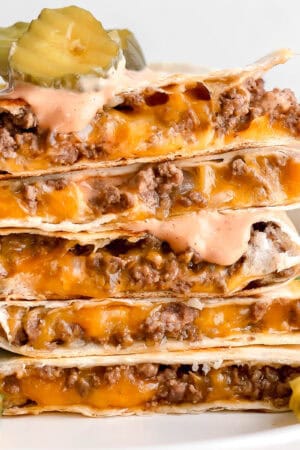 A stack of cheeseburger quesadillas with pickles and burger sauce on top and next to it.