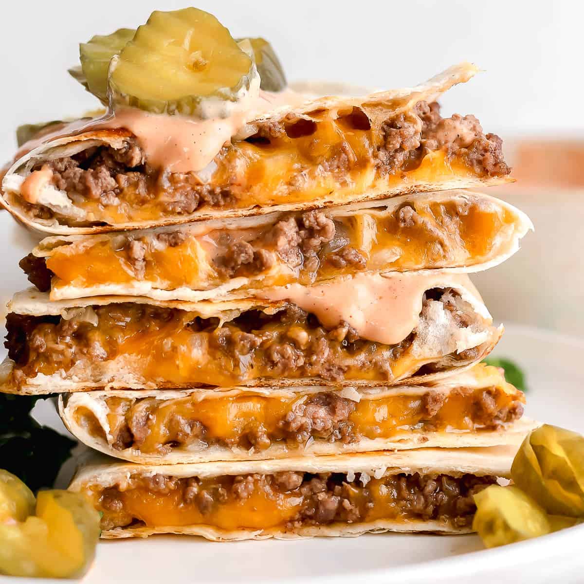 A stack of cheeseburger quesadillas with pickles and burger sauce on top and next to it.