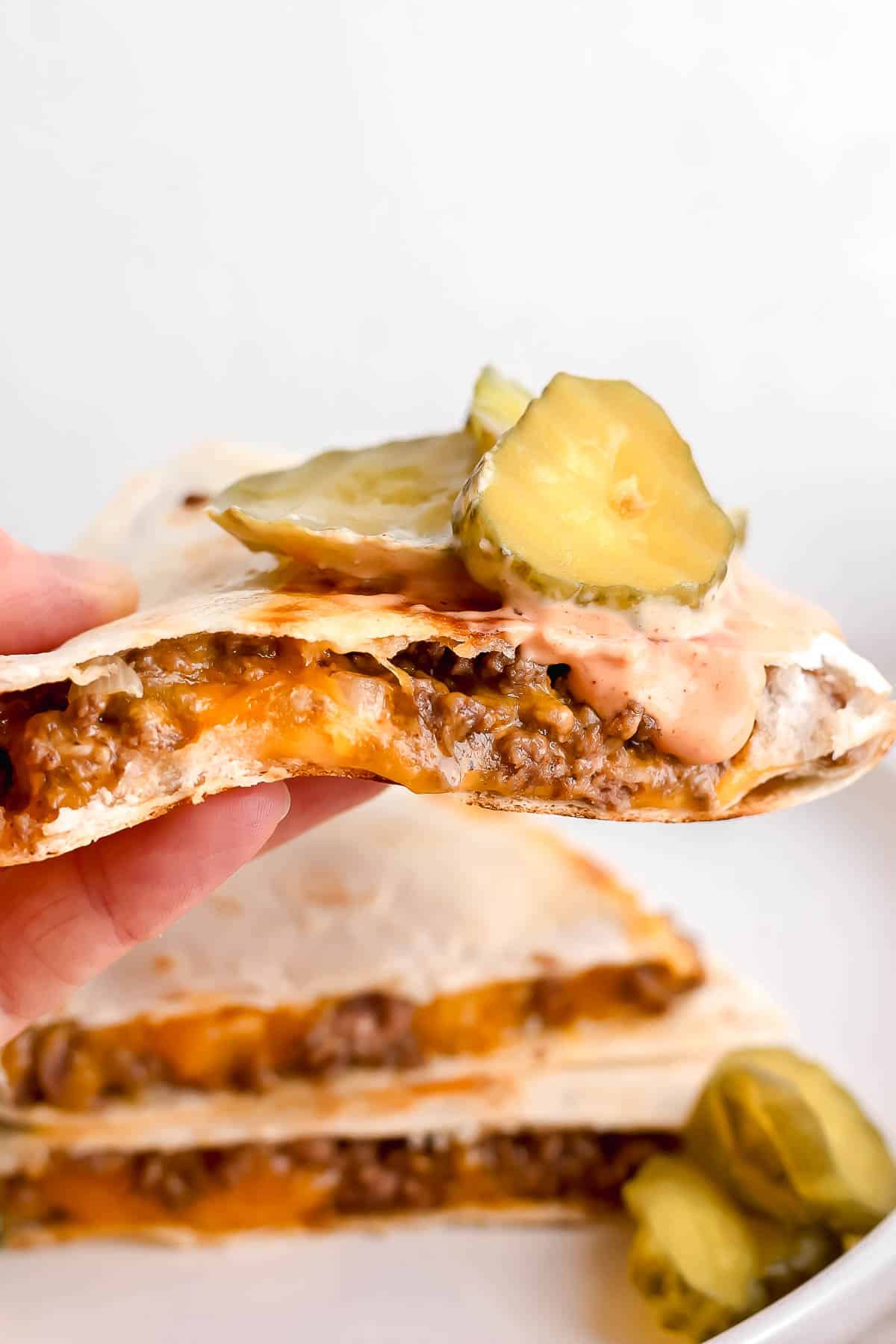 A hand holding a cheeseburger quesadilla topped with burger sauce and sliced pickles.