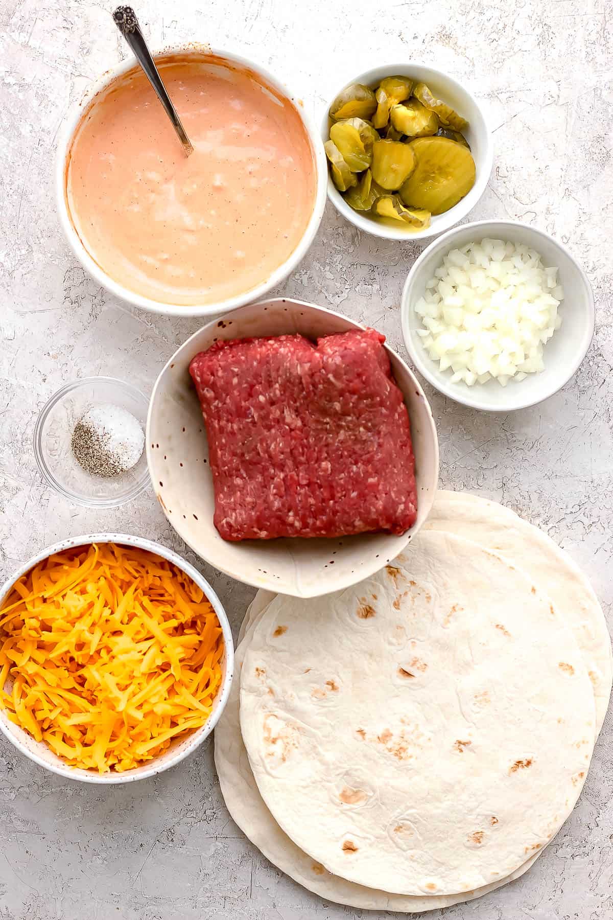 Individual bowls of burger sauce, sliced pickles, diced white onion, ground beef, salt & pepper, shredded cheese, and tortillas.