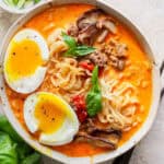 A bowl of coconut curry ramen with soft-boiled egg cut in half on top, crispy ground beef, fresh basil and mushrooms.