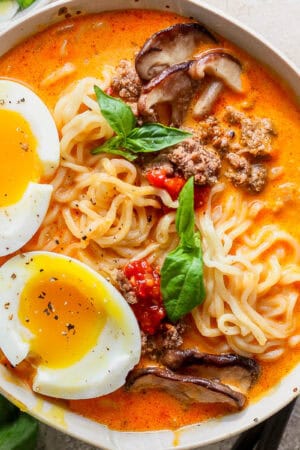 A bowl of coconut curry ramen with soft-boiled egg cut in half on top, crispy ground beef, fresh basil and mushrooms.