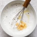 A bowl of Garlic White Sauce with a whisk sticking out and some garlic and pepper sitting on top.