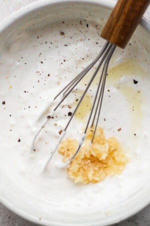 A bowl of Garlic White Sauce with a whisk sticking out and some garlic and pepper sitting on top.