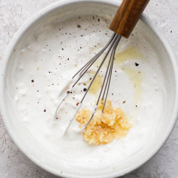 A bowl of Garlic White Sauce with a whisk sticking out and some garlic and pepper sitting on top.