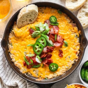 A small cast iron skillet filled with a warm jalapeno popper dip topped with bacon, green onion and sliced jalapeno with a piece of bread sticking out.