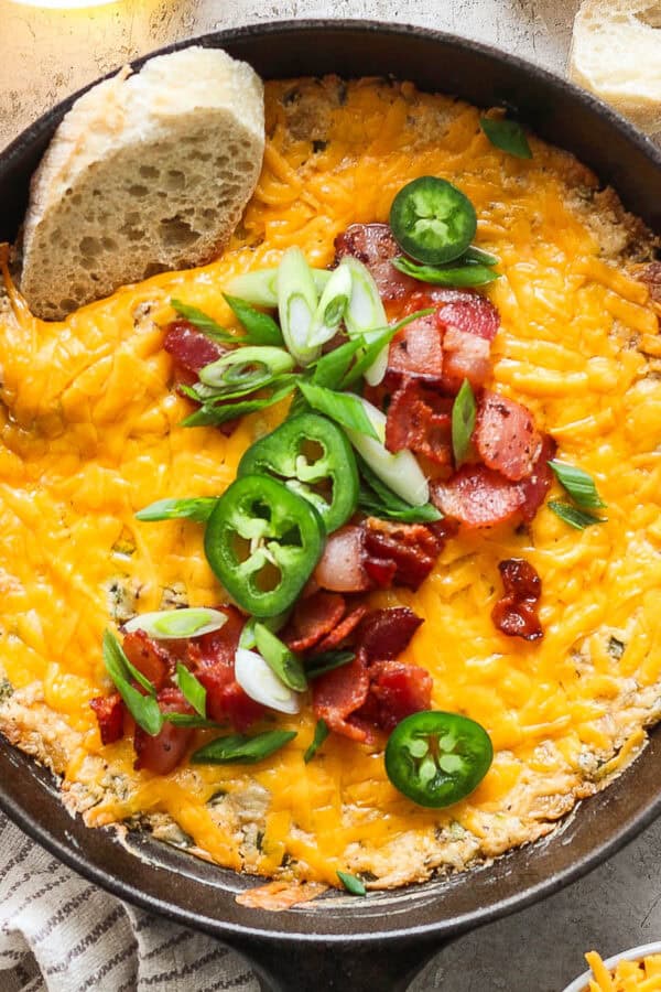 A small cast iron skillet filled with a warm jalapeno popper dip topped with bacon, green onion and sliced jalapeno with a piece of bread sticking out.