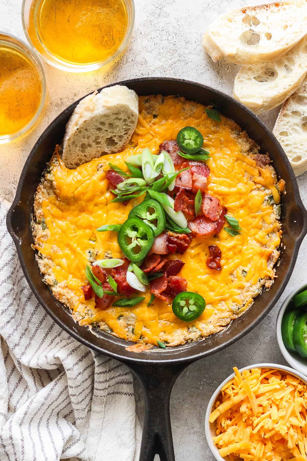 Jalapeno popper dip in a skillet surrounded by a few glasses of beer and sliced bread.