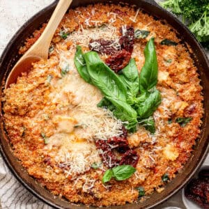 A cast iron skillet filled with marry me chicken quinoa bake with fresh basil, shredded parmesan cheese and sun-dried tomatoes on top and a wooden spoon sticking out.