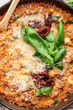 A cast iron skillet filled with marry me chicken quinoa bake with fresh basil, shredded parmesan cheese and sun-dried tomatoes on top and a wooden spoon sticking out.