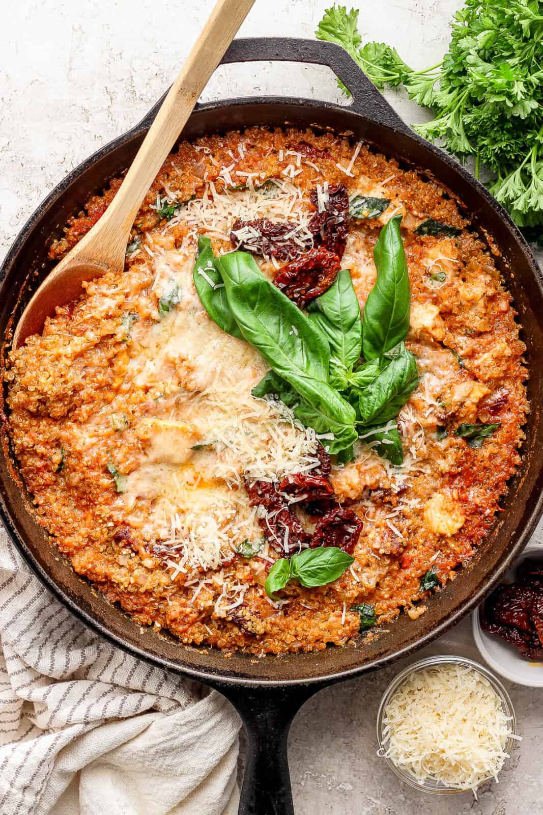A cast iron skillet of marry me chicken quinoa with a wooden spoon sticking out the side and fresh basil on top.