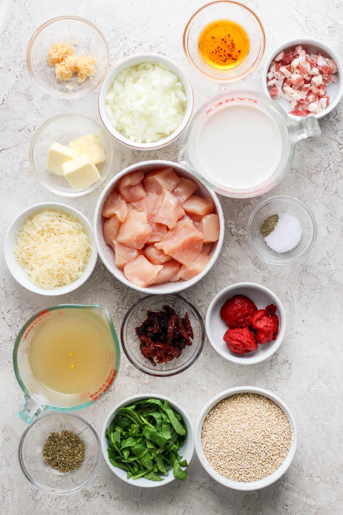 Individual bowls of minced garlic, diced white onion, butter, pancetta, salt and pepper, cut up chicken chunks, half & half, tomato paste, chicken stock, quinoa, spinach, sun-dried tomoatoes, sun-dried tomato oil, italian seasoning, and parmesan cheese.