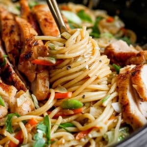 A cast iron skillet filled with homemade chicken chow mein with sliced chicken thighs and someone twirling pasta on a fork.