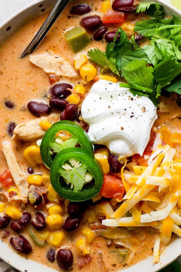 A bowl of creamy chicken taco soup with sliced jalapenos, sour cream, shredded cheese and fresh cilantro on top with a spoon sticking out.