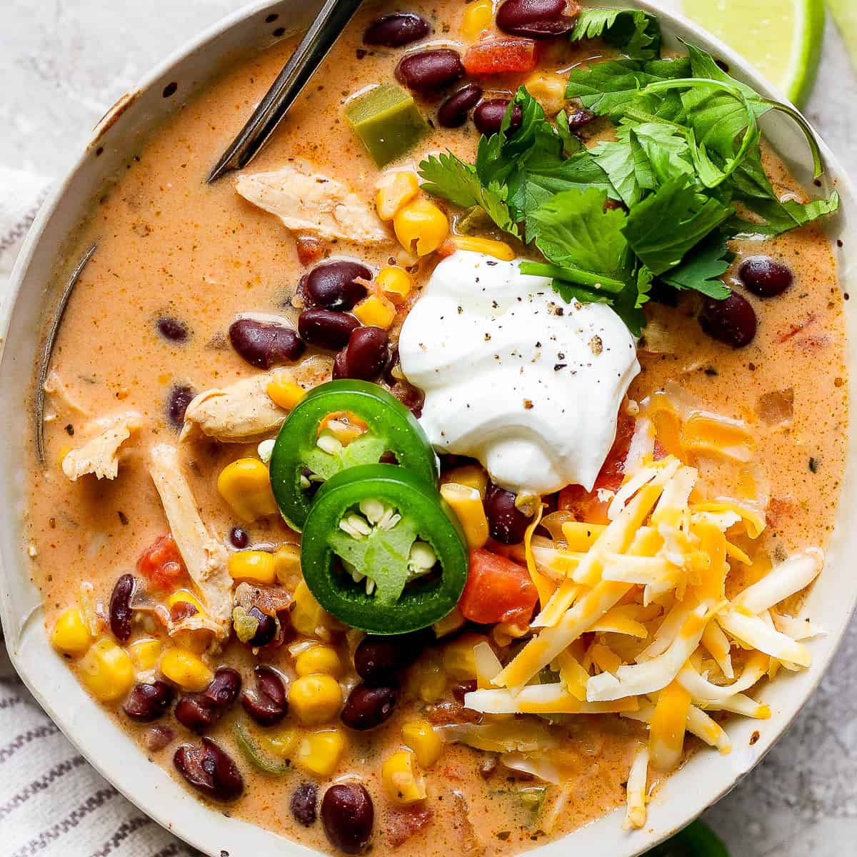 A bowl of creamy chicken taco soup with sliced jalapenos, sour cream, shredded cheese and fresh cilantro on top with a spoon sticking out.