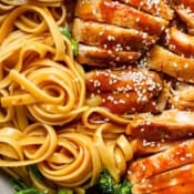 Close up shot of twirled teriyaki noodles with sliced chicken thighs next to it and some broccoli with sesame seeds on top.