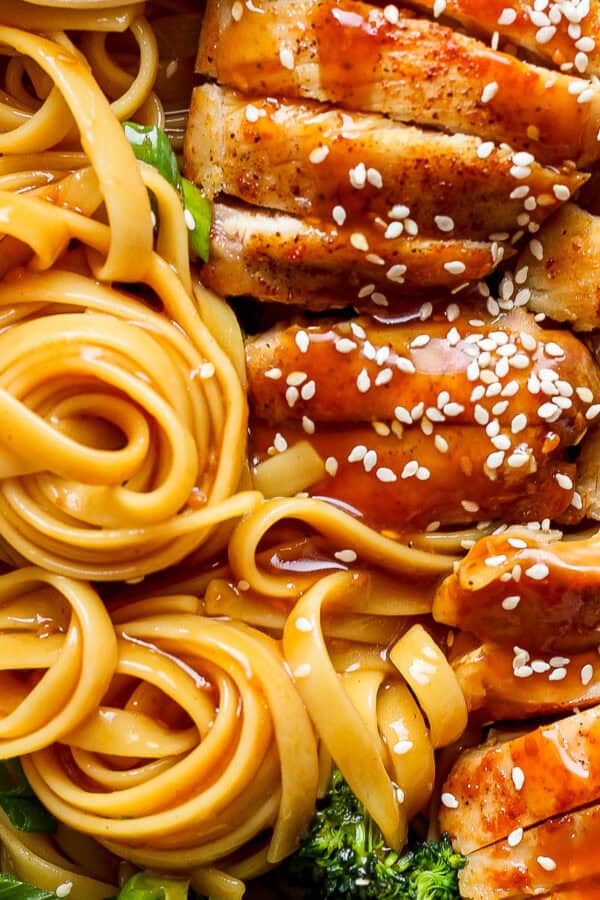 Close up shot of twirled teriyaki noodles with sliced chicken thighs next to it and some broccoli with sesame seeds on top.