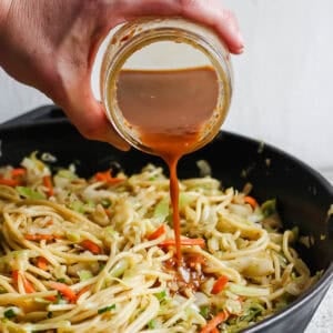 Someone pouring homemade chow mein sauce over a skillet filled with chicken chow mein.