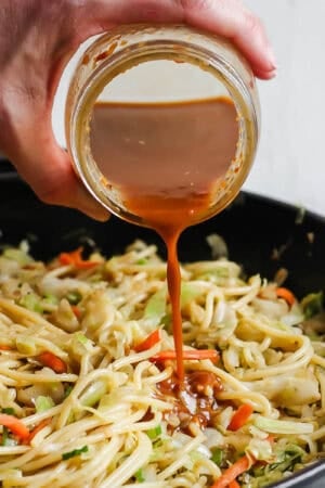 Someone pouring homemade chow mein sauce over a skillet filled with chicken chow mein.