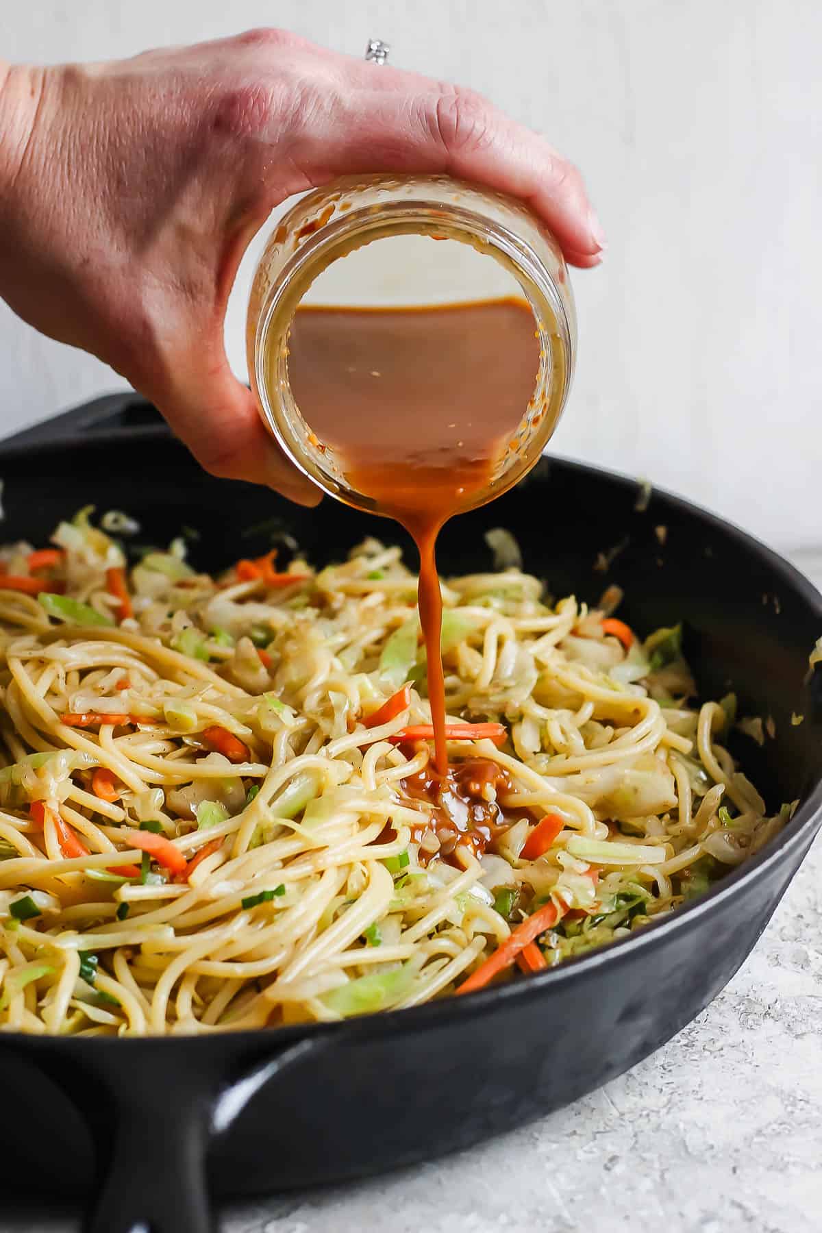 A hand pouring a jar of chicken chow mein sauce into a skillet of chicken chow mein.