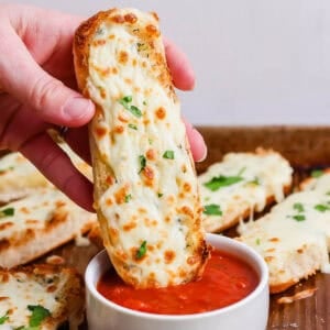 Someone holding an Italian Dunker while dipping it into some marinara sauce with a baking sheet filled with more Italian Dunkers underneath.