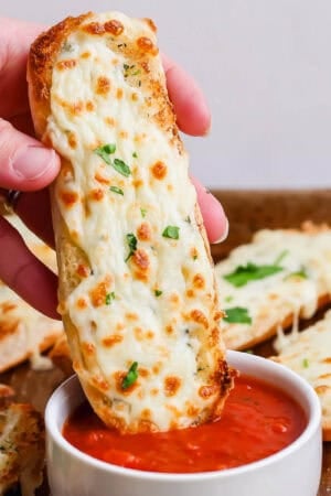 Someone holding an Italian Dunker while dipping it into some marinara sauce with a baking sheet filled with more Italian Dunkers underneath.