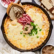 A small cast iron skillet filled with a reuben dip and topped with sauerkraut, shredded corned beef and parsley with 3 pieces of marbled rye sticking out.