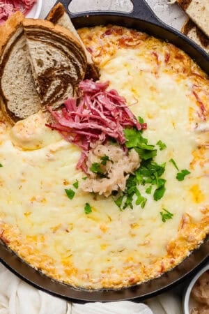 A small cast iron skillet filled with a reuben dip and topped with sauerkraut, shredded corned beef and parsley with 3 pieces of marbled rye sticking out.