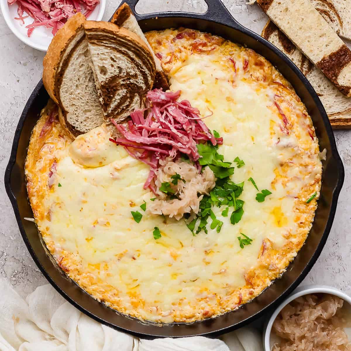 A small cast iron skillet filled with a reuben dip and topped with sauerkraut, shredded corned beef and parsley with 3 pieces of marbled rye sticking out.