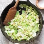 A large cast iron skillet filled with sauteed cabbage with a wooden spatula sticking out and a salt pinch bowl next to it.