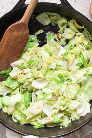 A large cast iron skillet filled with sauteed cabbage with a wooden spatula sticking out and a salt pinch bowl next to it.