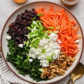 A bowl of carrot salad with raisins, kale, green onions, walnuts and goat cheese crumbles.