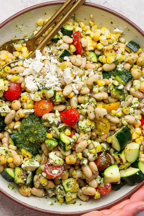 A big bowl of dense bean summer salad with two spoon sticking out and two hands holding the bowl.