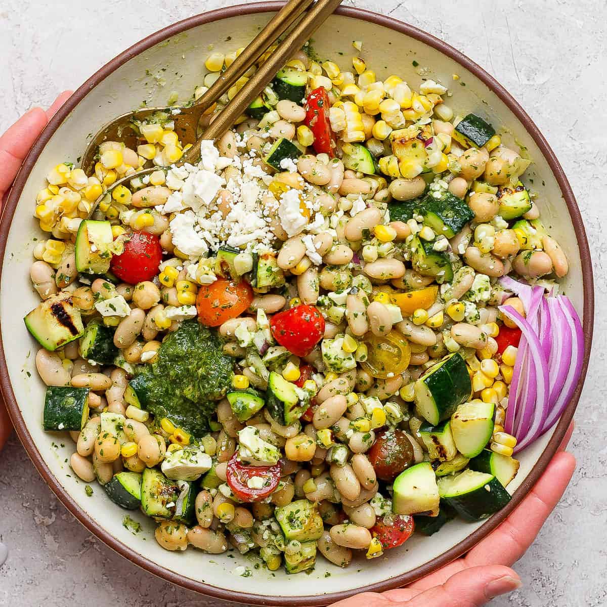 A big bowl of dense bean summer salad with two spoon sticking out and two hands holding the bowl.