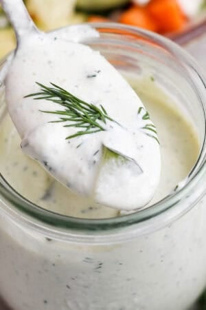A jar of homemade pickle ranch dressing with a spoon lifting some out with a piece of dill on top.