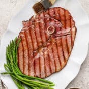 A platter with a grilled ham steak on top with steamed asparagus and a brush with glaze on it.