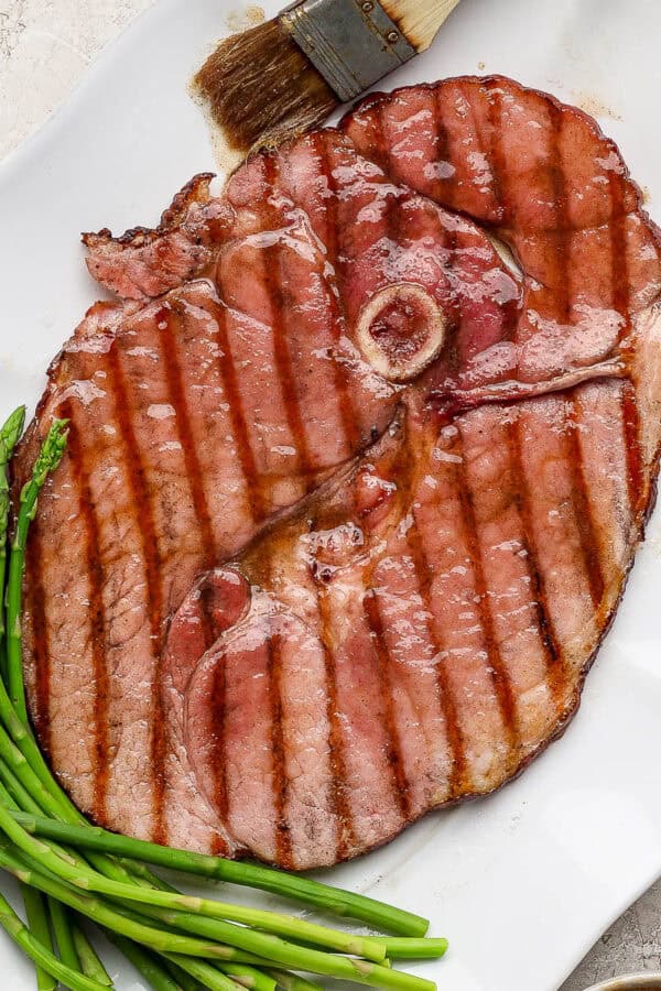 A platter with a grilled ham steak on top with steamed asparagus and a brush with glaze on it.