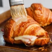A melted ham and cheese croissant on a baking sheet with a brush adding some honey mustard sauce on top.